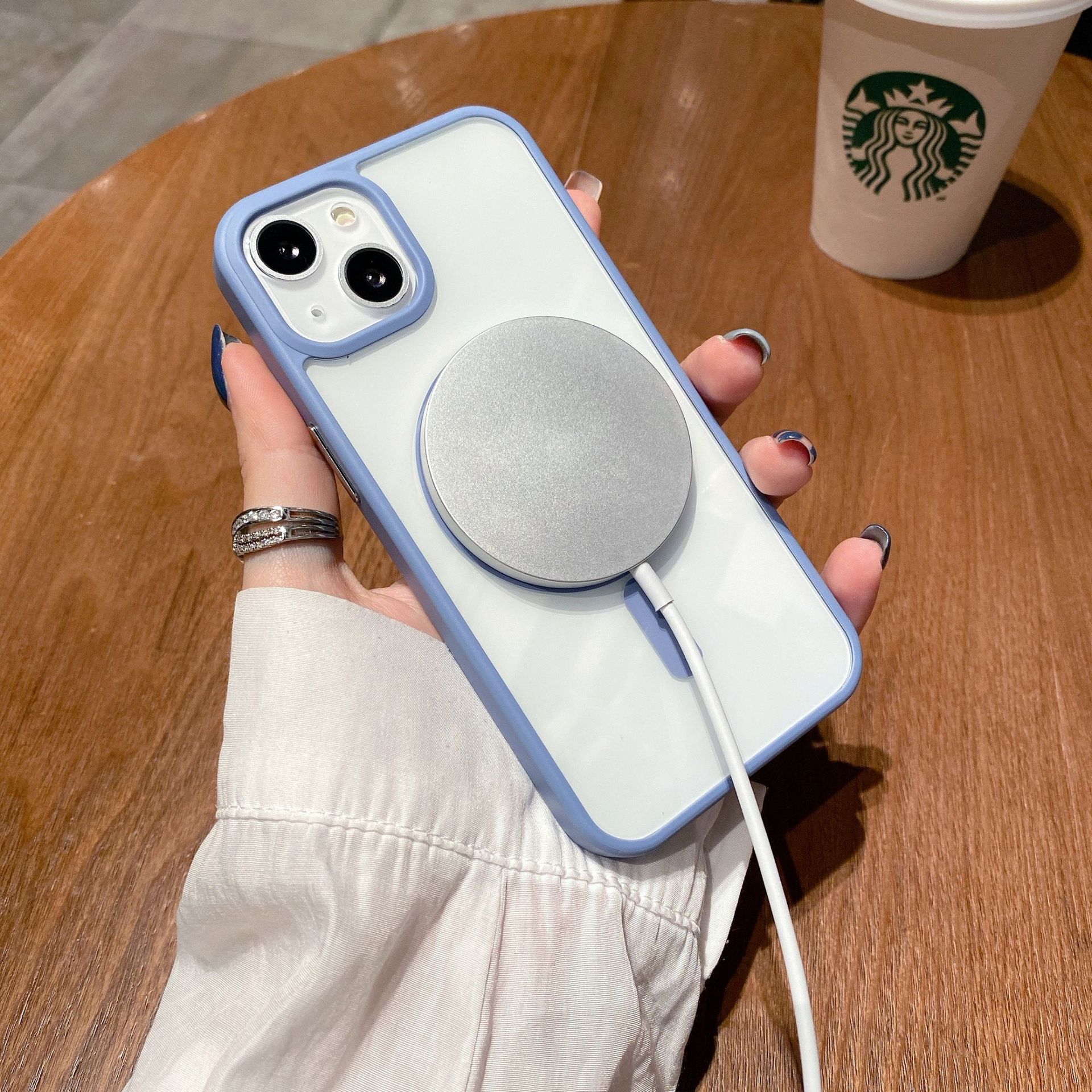 Phone Case transparente de acrílico con carga MagSafe y cable conectado, sosteniendo en mano junto a una bebida de Starbucks.